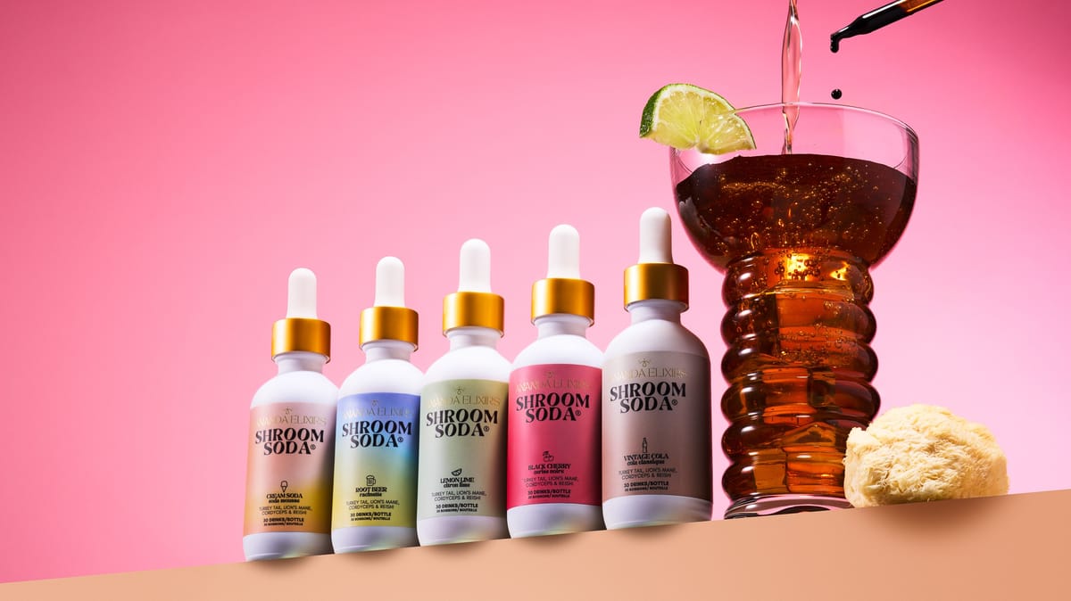 Five Shroom Soda bottles in a row beside a glass of amber soda with lime, against a bright pink backdrop