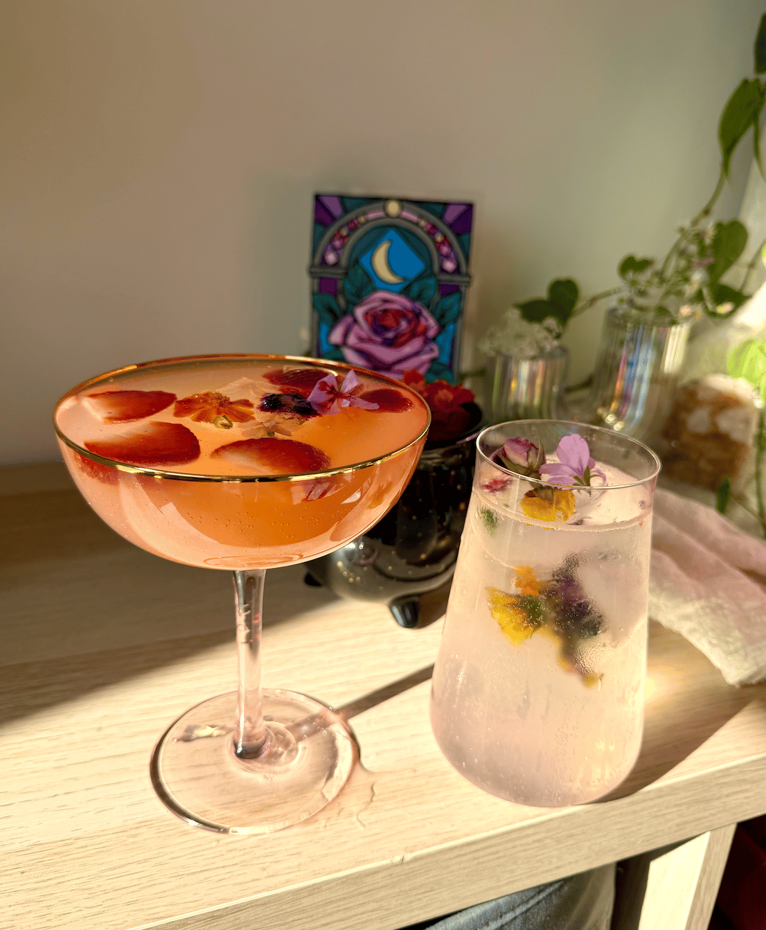 Two pink cocktails garnished with sliced strawberries and edible flowers sit on a table.