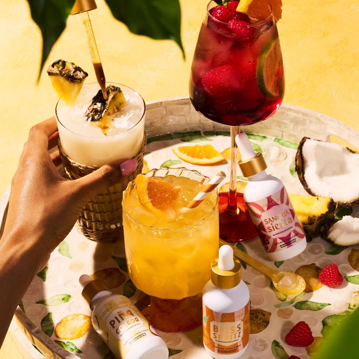 A hand holds a tropical cocktail on a tray surrounded by drinks and dropper bottles.